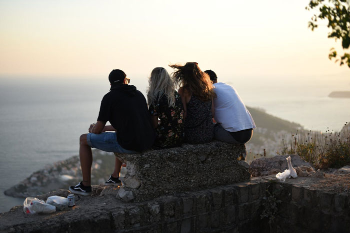 Four friends sitting on a stone bench overlooking the sea, illustrating the couple hosting an old friend with regret. Four friends sitting on a stone bench overlooking the sea, illustrating the couple hosting an old friend with regret.