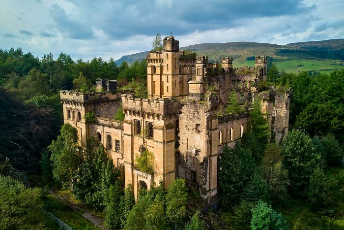 Abandoned castle overtaken by trees and vegetation, surrounded by dense forest and hills under a cloudy sky.