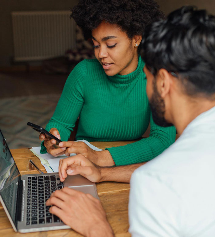 Couple discussing postpartum trip plans while using laptop and phone, focusing on decision involving pregnant wife and child.