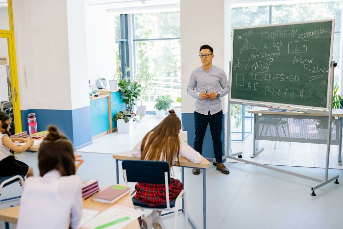 Teacher explaining math questions on a chalkboard to students in a bright classroom for grade levels 1 to 12.