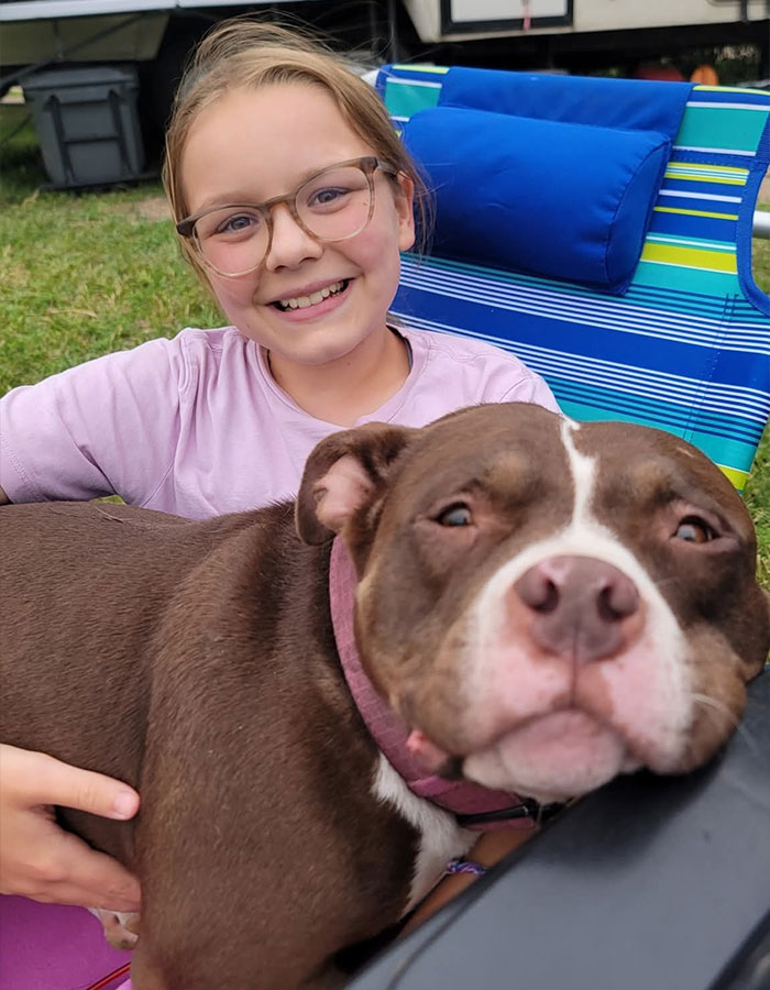Smiling young girl in glasses sitting outdoors holding a large brown dog, capturing a heartfelt family moment before explosion tragedy.