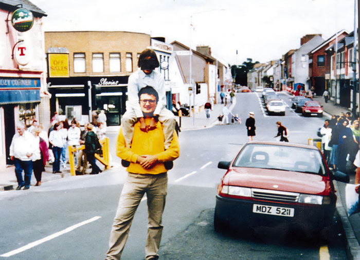 Man carrying child on shoulders near a street with a car and crowd, one of the photos taken before disaster moments.