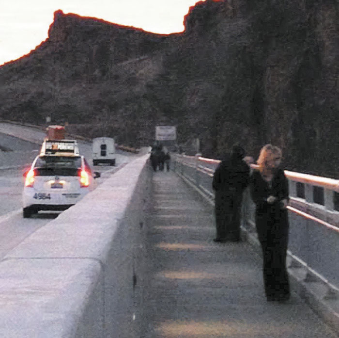 Photo taken before disaster showing people on a bridge with a parked car and mountains in the background at dusk.
