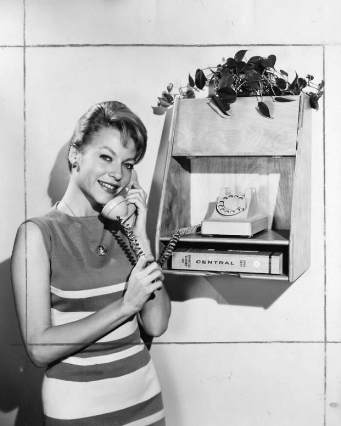 LA woman in 1962 smiling while using a vintage rotary phone in a stylish striped dress near a wall-mounted shelf.