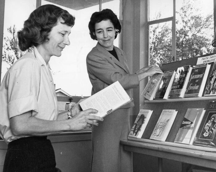 Two LA women in 1962 browsing books in a sunlit room, enjoying a peaceful and engaging moment together.