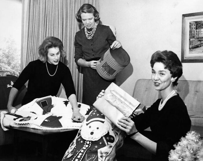 Three LA women in 1962, dressed in vintage attire, engaging in festive holiday decorating indoors.