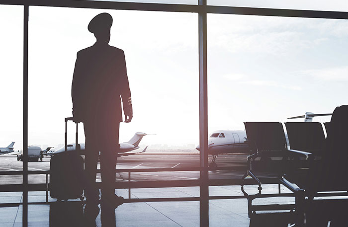 Silhouette of a pilot in uniform with suitcase standing in an airport terminal near airplanes at sunrise.