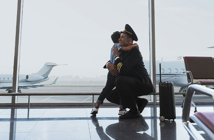 Pilot in uniform hugging child at airport terminal with private jets outside, reflecting on terrifying pilot situations.