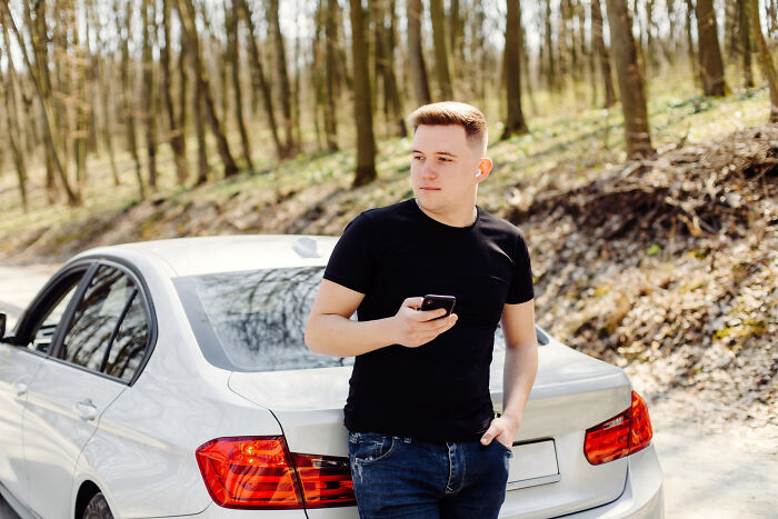 Teenager standing by a white car in a forest area, holding a phone, representing police concerns about illegal kids and teenagers.