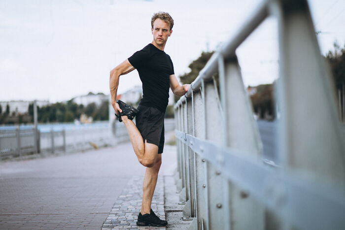 Teenager in black sportswear stretching leg outdoors near railing during morning workout session.