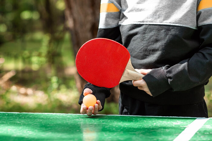 Teenager holding a red ping pong paddle and orange ball, preparing to serve on a green outdoor table tennis table.