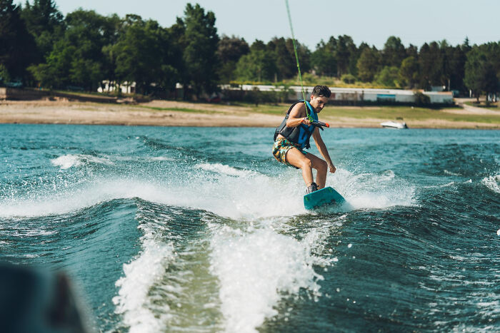 Teenager wakeboarding on a lake near the shore with trees, no police or illegal activity visible in the scene.