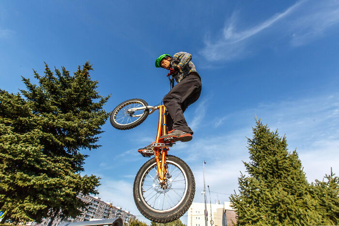 Teenager wearing a green helmet performing a bicycle stunt outdoors with trees and blue sky in the background.