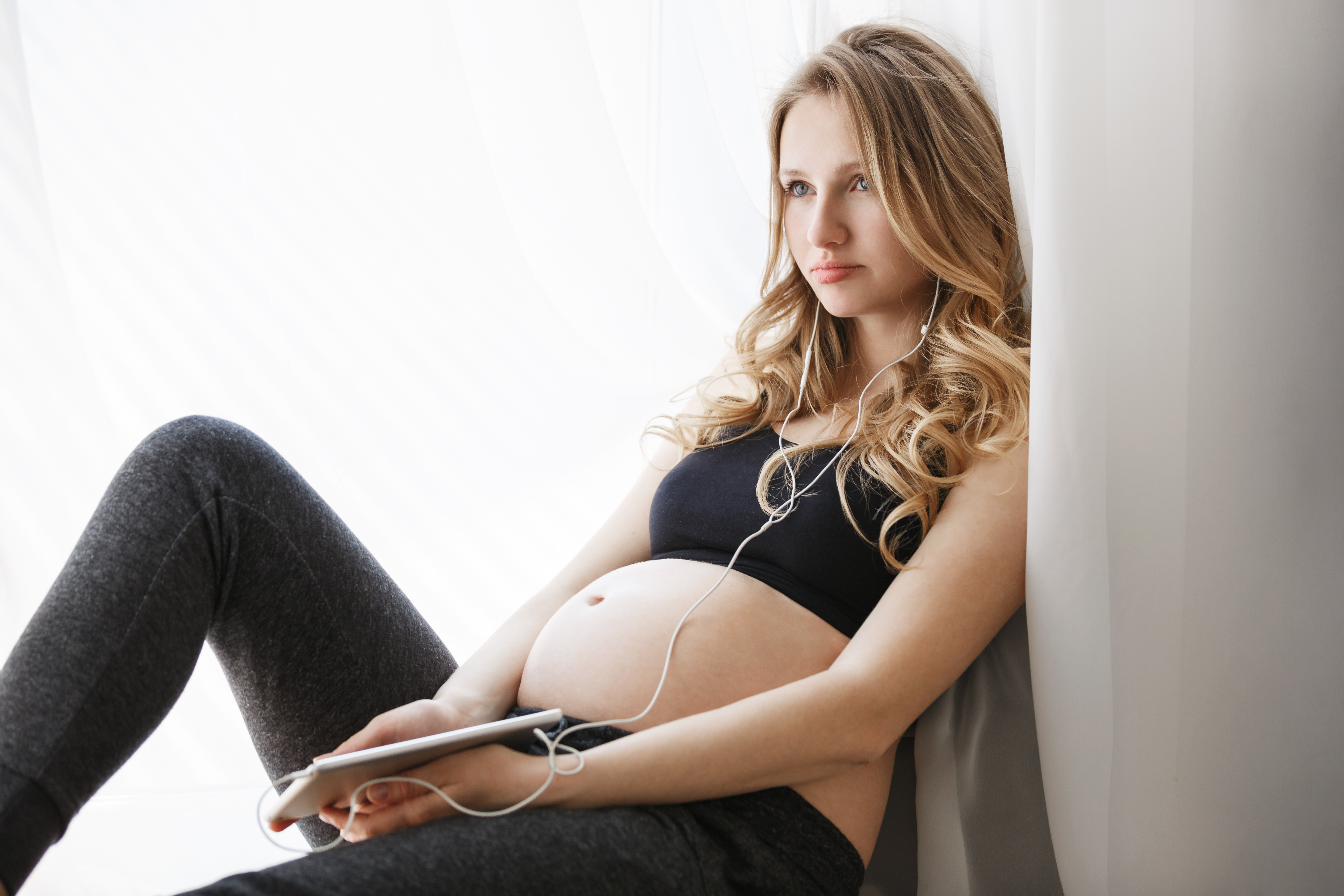 Pregnant young woman sitting by window, listening to music, reflecting on estranged dad and reconnecting with daughter story.