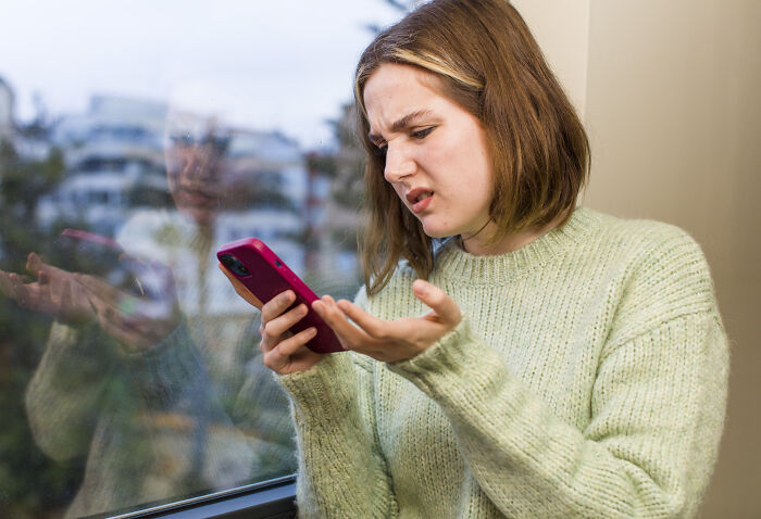 Young woman shocked while reading secrets about hidden families and mob money on her phone by a window.