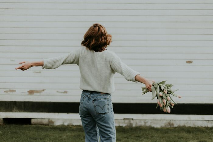 Person holding tulips with arms outstretched standing outside against a white wall, illustrating gender-based double standards.