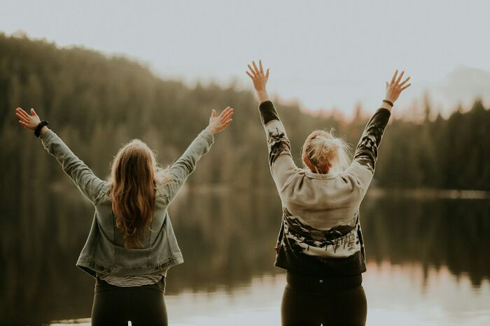 Two people by the lake raising hands joyfully, representing good news and positivity in an outdoor nature setting.