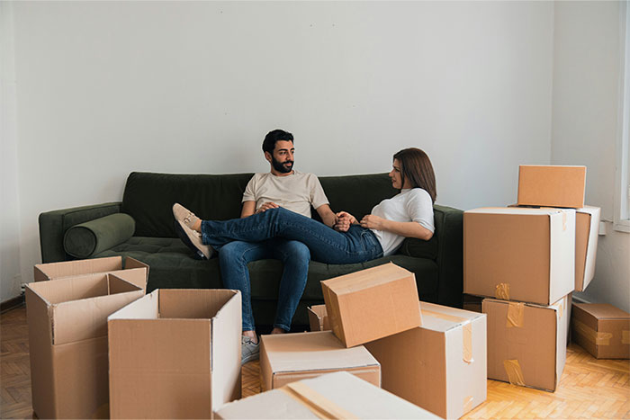 Couple sitting on a green couch surrounded by cardboard boxes, appearing deep in conversation about a psychiatrist visit. Couple sitting on a green couch surrounded by cardboard boxes, appearing deep in conversation about a psychiatrist visit.