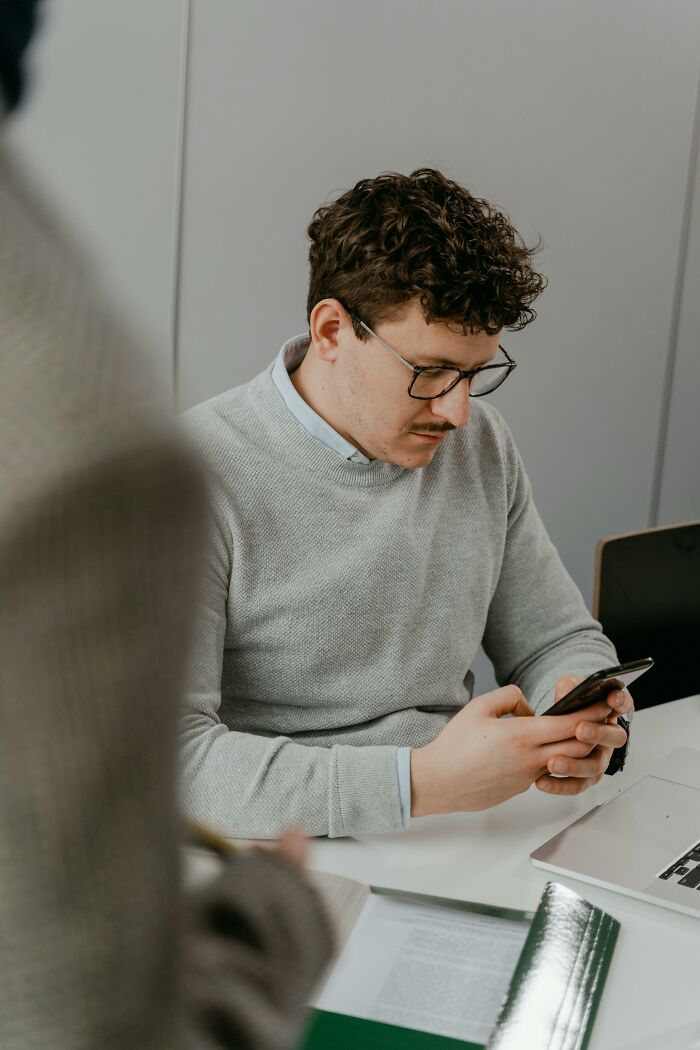 Man with glasses using smartphone at desk, representing people exposing performative behaviors in society.