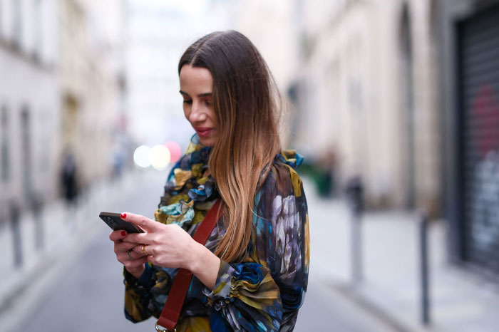 Young woman in a patterned blouse texting on her phone outdoors, capturing mood-based questions to ask your crush.