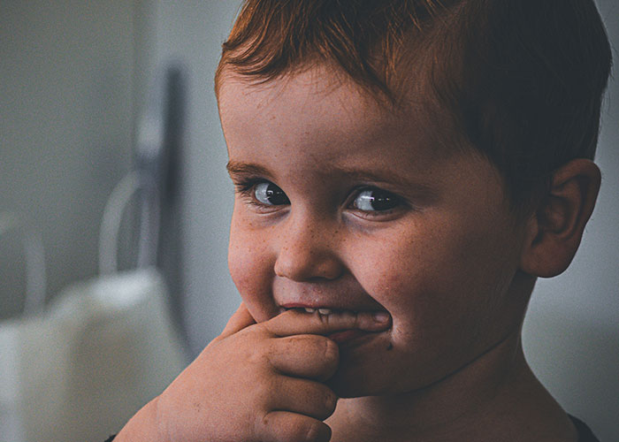 Close-up of a young child with red hair and freckles, showing quick reactions by biting fingers in a thoughtful pose.