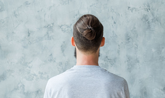 Man with a bun hairstyle wearing a gray shirt standing against a textured background, reflecting rage room customers needing therapy.