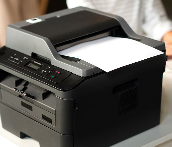 Black office printer on a white desk with a sheet of paper partially printed, related to rage room customers needing therapy.