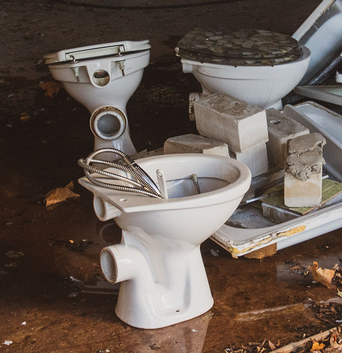 Three broken toilets and scattered debris in a rage room setting with bricks and metal parts on wet floor.
