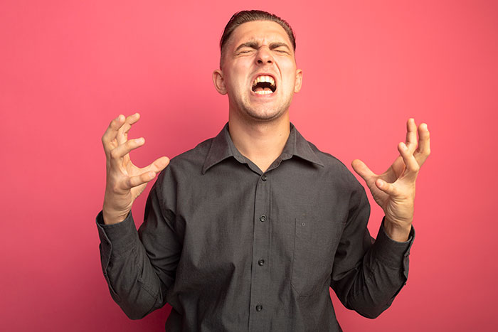 Young man expressing rage with clenched hands and eyes closed against a pink background, illustrating rage room customers needing therapy