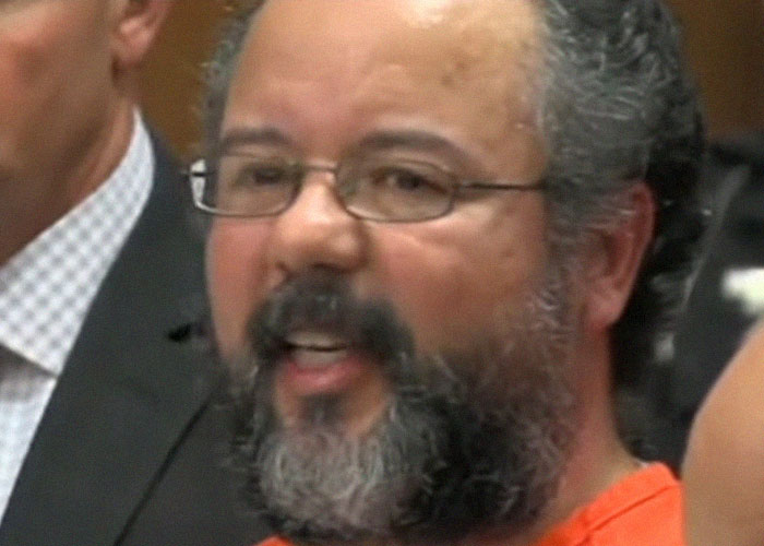 Man with glasses and beard speaking in a crowded setting, capturing a moment of real-life terror and paranoia.