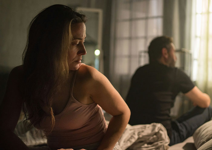 Woman looking uneasy on a bed while a man sits turned away, illustrating behaviors that might make women feel unsafe.