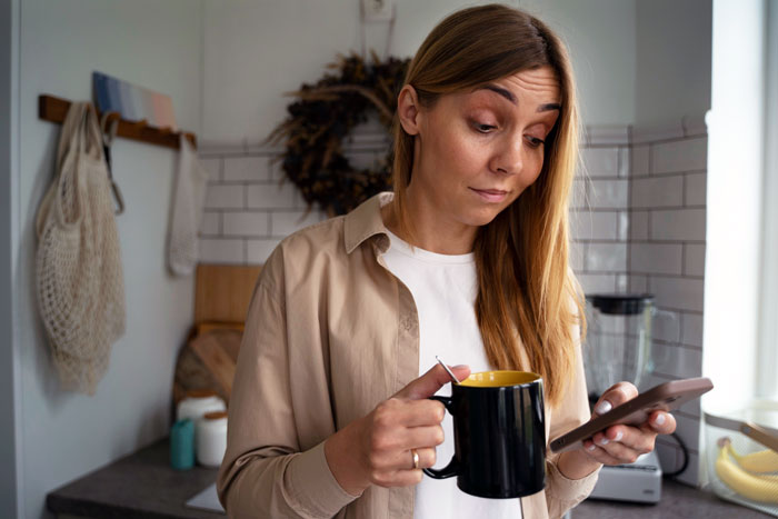 Young woman in kitchen holding a black mug and phone, showing hesitation about refuse female friend use shampoo advice.