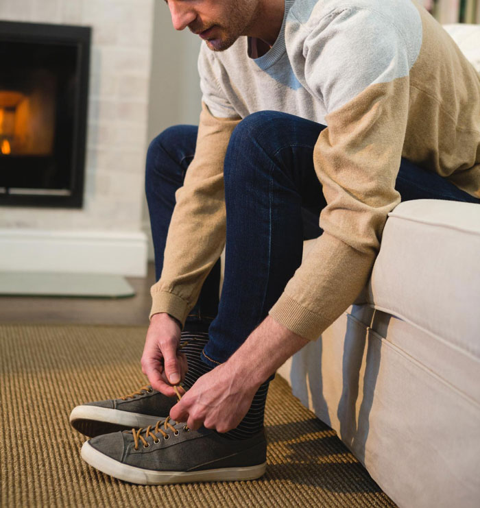 Man sitting on couch tying shoes indoors, illustrating guy with medical condition not removing shoes in friend's house.