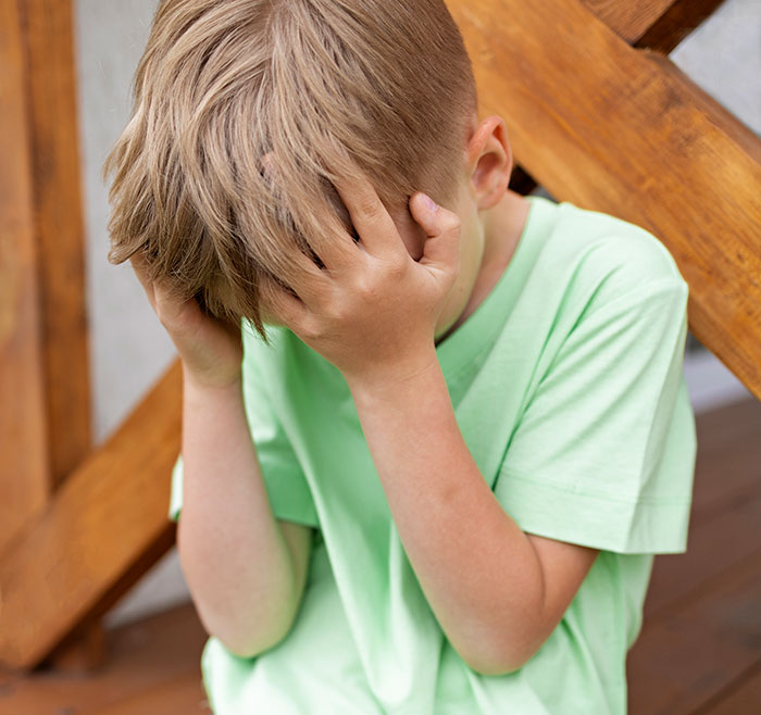 Young boy in green shirt covering his face, depicting emotional distress related to growing up with a psychopath experience.