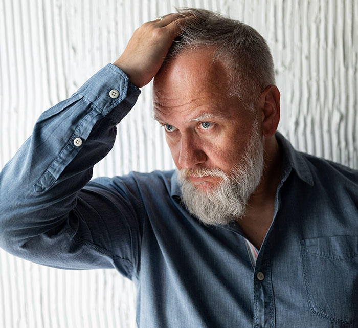 Middle-aged man with gray beard and blue shirt, looking worried while touching his head, reflecting on growing up with a psychopath.