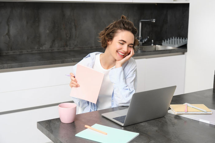 Young woman smiling at laptop while holding notebook in modern kitchen, representing tenant and landlord conflict story.