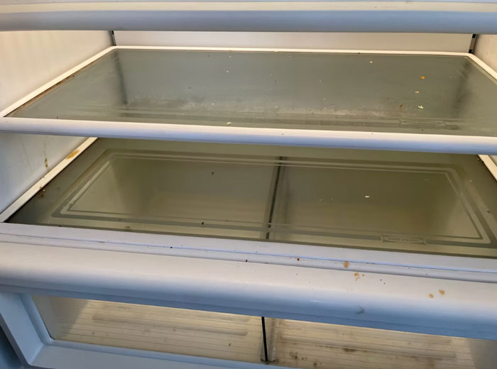 Empty dirty refrigerator shelves showing damage inside an apartment after tenant quits paying rent and causes wreckage.