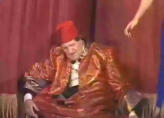 Man in an ornate red outfit and fez hat seated against a red curtain, part of videos that gain meaning with more context. - 19