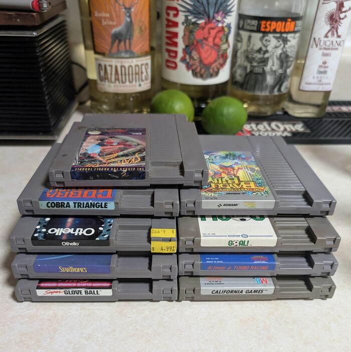 Stack of retro gaming cartridges from the 80s and 90s displayed with limes and bottles in the background.
