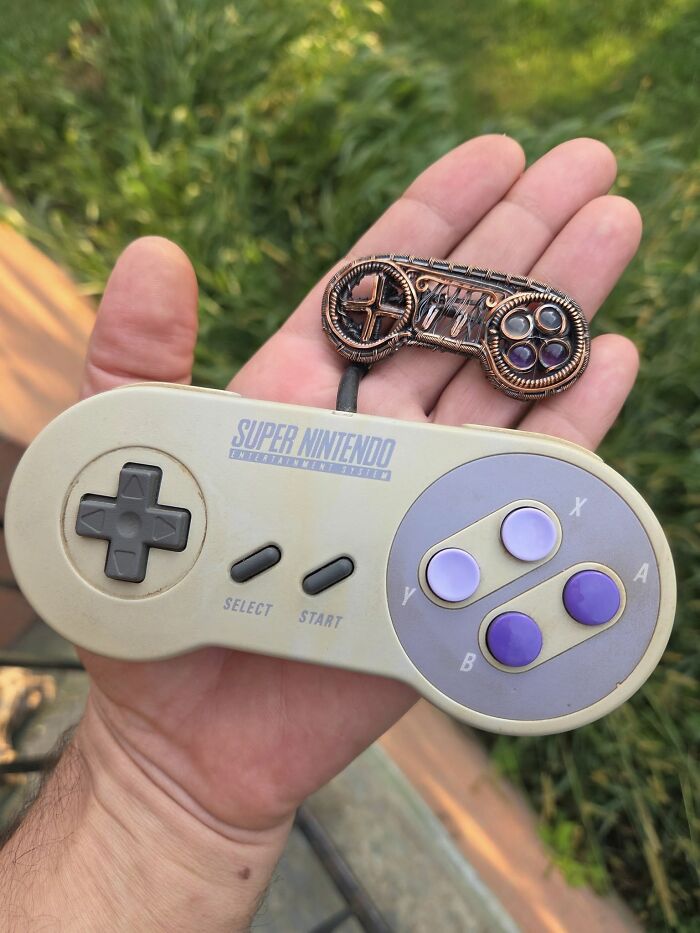 Hand holding a vintage Super Nintendo controller and a wire-wrapped miniature retro gaming controller outdoors.