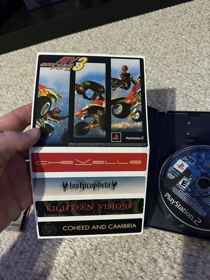 Hand holding a PlayStation 2 ATV Offroad Fury 3 game insert with band stickers, next to an open game case on carpet.