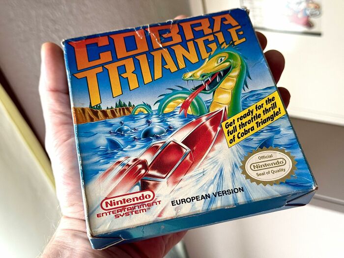 Hand holding a retro gaming Cobra Triangle Nintendo Entertainment System box with vintage 80s game artwork.