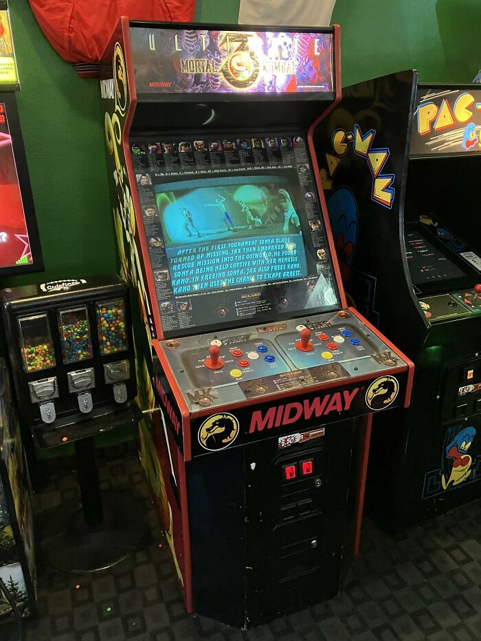 Retro gaming arcade machine featuring Mortal Kombat by Midway, next to a Pac-Man arcade and colorful gumball dispensers.