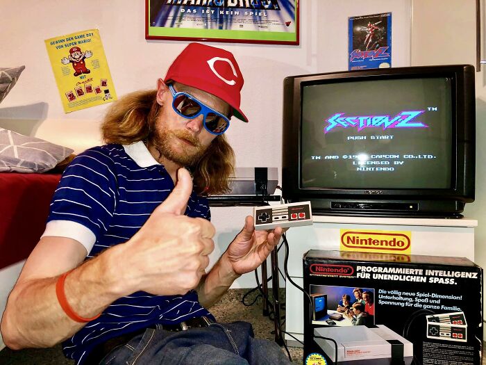 Man in a red cap and sunglasses playing retro Nintendo game, showing thumbs up in a nostalgic gaming setup.