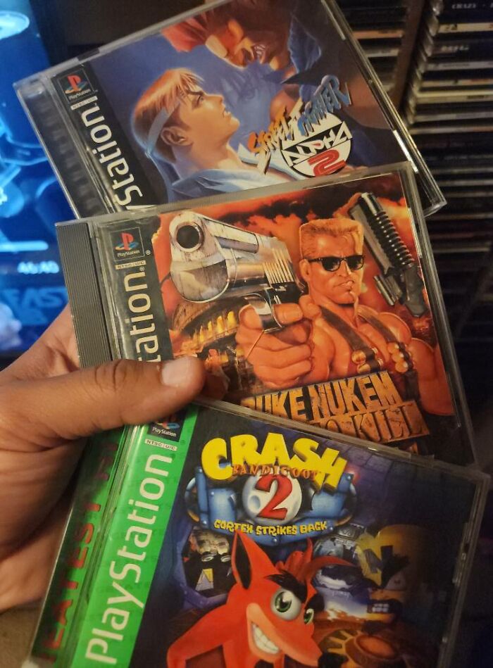 Hand holding retro PlayStation game cases including Street Fighter, Duke Nukem, and Crash Bandicoot for gaming nostalgia.