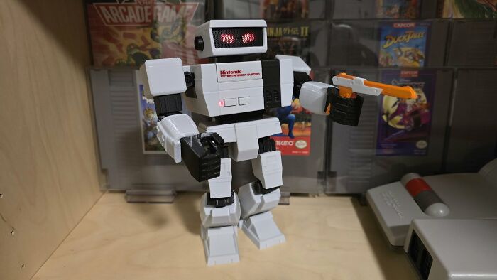 Retro gaming robot figure holding an orange toy gun with classic video game cartridges displayed in the background.