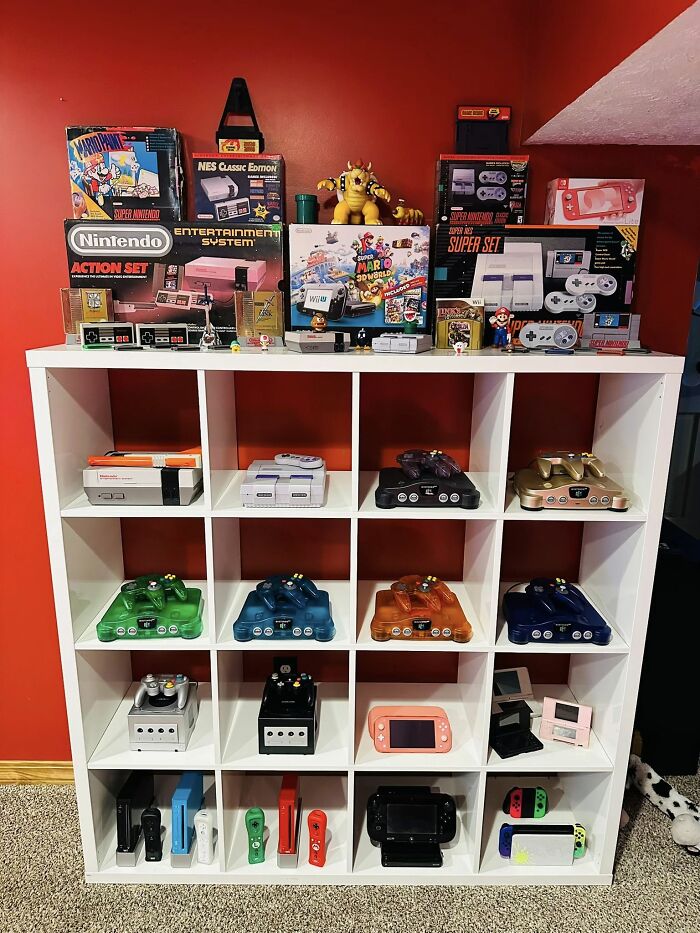 Collection of retro gaming consoles and accessories displayed on white shelves against a red wall, evoking nostalgia.