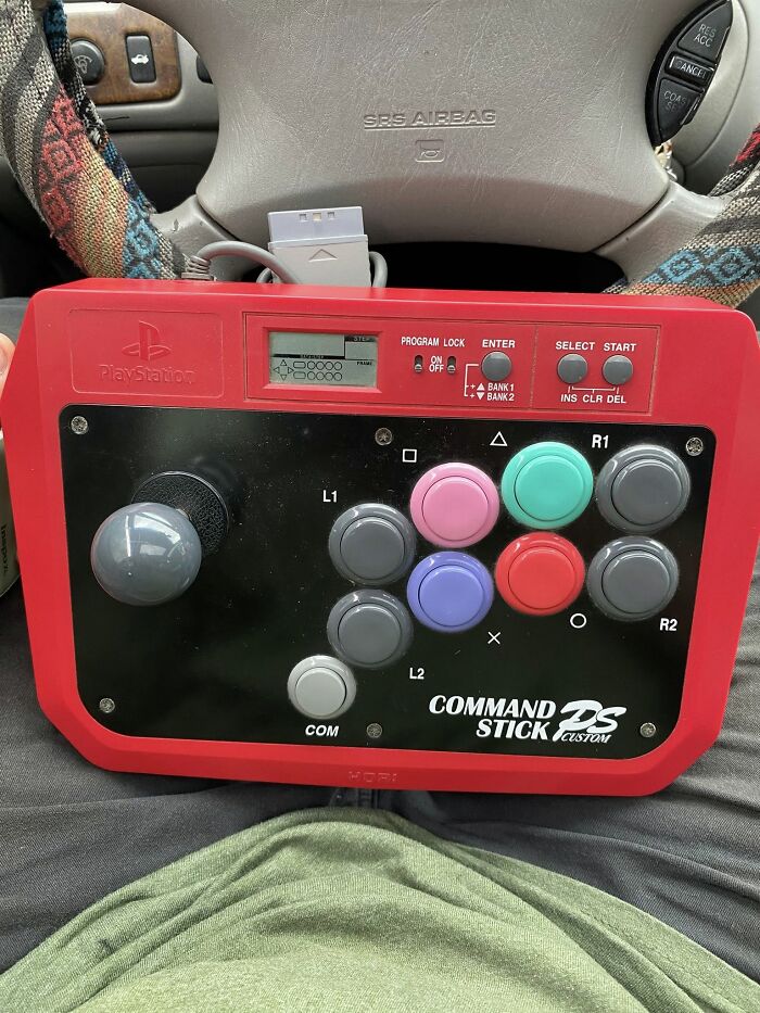 Retro gaming controller with colorful buttons resting on a person's lap inside a car, evoking nostalgia.