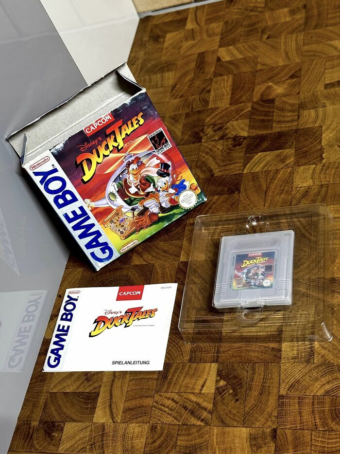 Retro gaming nostalgia with Disney DuckTales Game Boy cartridge, manual, and original packaging on wooden surface.