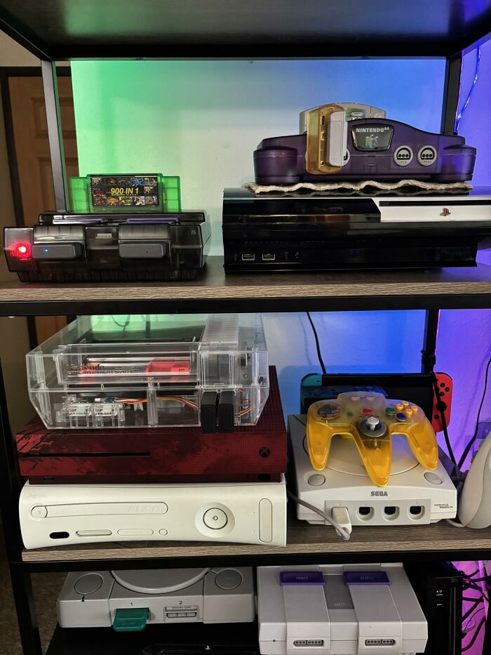 Retro gaming consoles including Nintendo 64, PlayStation, Xbox, Sega Dreamcast, and retro controllers on wooden shelves.
